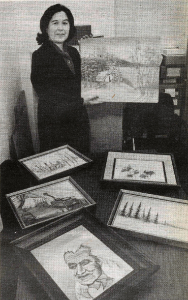 Black and white photograph of Elder G. Jean Crane standing in a room and holding up one of her paintings, which depicts a winter landscape with a snow-covered cabin and trees. She is dressed in dark professional attire and smiling slightly. On the table in front of her are six framed artworks, including images of wildlife, natural scenery, and a portrait of Chief Dan George.