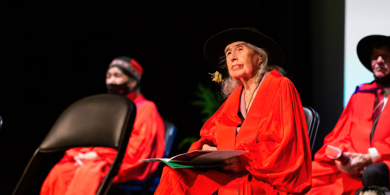 Elder G. Jean Crane sits on stage wearing a bright red academic gown and a black ceremonial hat, holding a convocation program in her lap. She gazes upward thoughtfully. Other individuals in similar academic regalia are seated nearby, slightly out of focus, against a dark background.