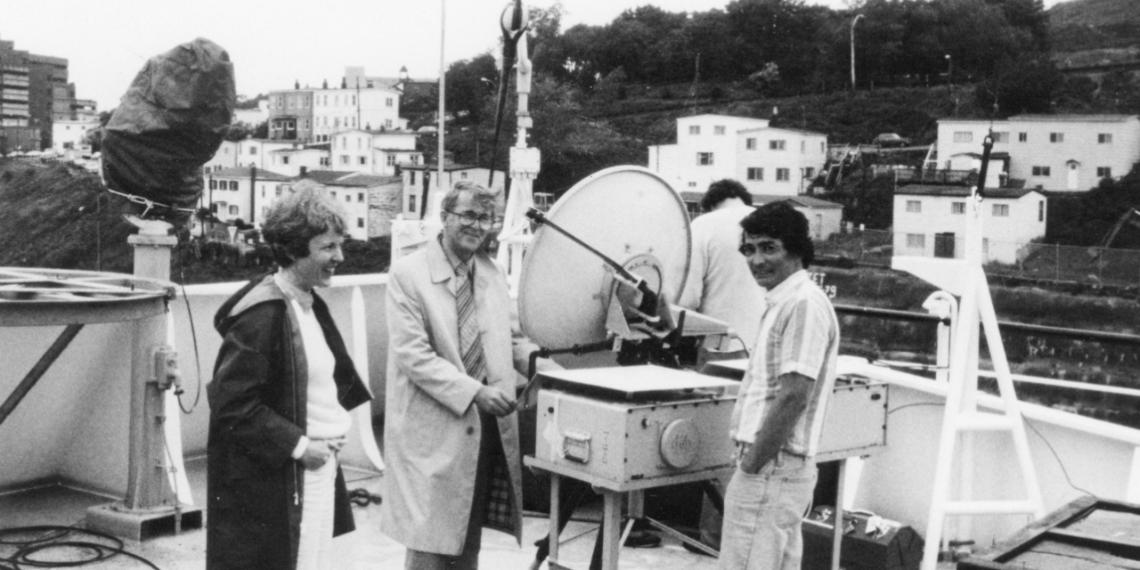 A black and white photograph showing four people standing near a small satellite dish and other pieces of equipment. Dr. Max House appears in the middle smiling at the camera a wearing a beige trench coat. In the background are Newfoundland-style salt-box house on a hillside.